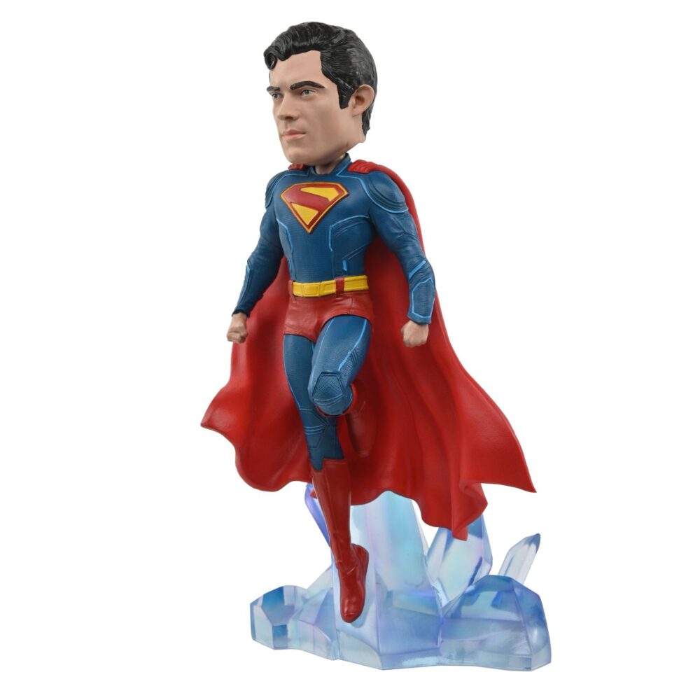 Superman 2025 dc universe head knocker. Neca presenta la figura de superman basada en el protagonista de la película homónima de 2025. Se muestra sobre una base translúcida de cristales de la fortaleza de la soledad, con su clásico traje azul y capa roja. Mide 21,5 cm, está fabricada con resina y pintada a mano con gran nivel de detalle. Cuenta con un mecanismo de resorte que permite el característico movimiento de cabeza de las figuras de la colección head knocker.