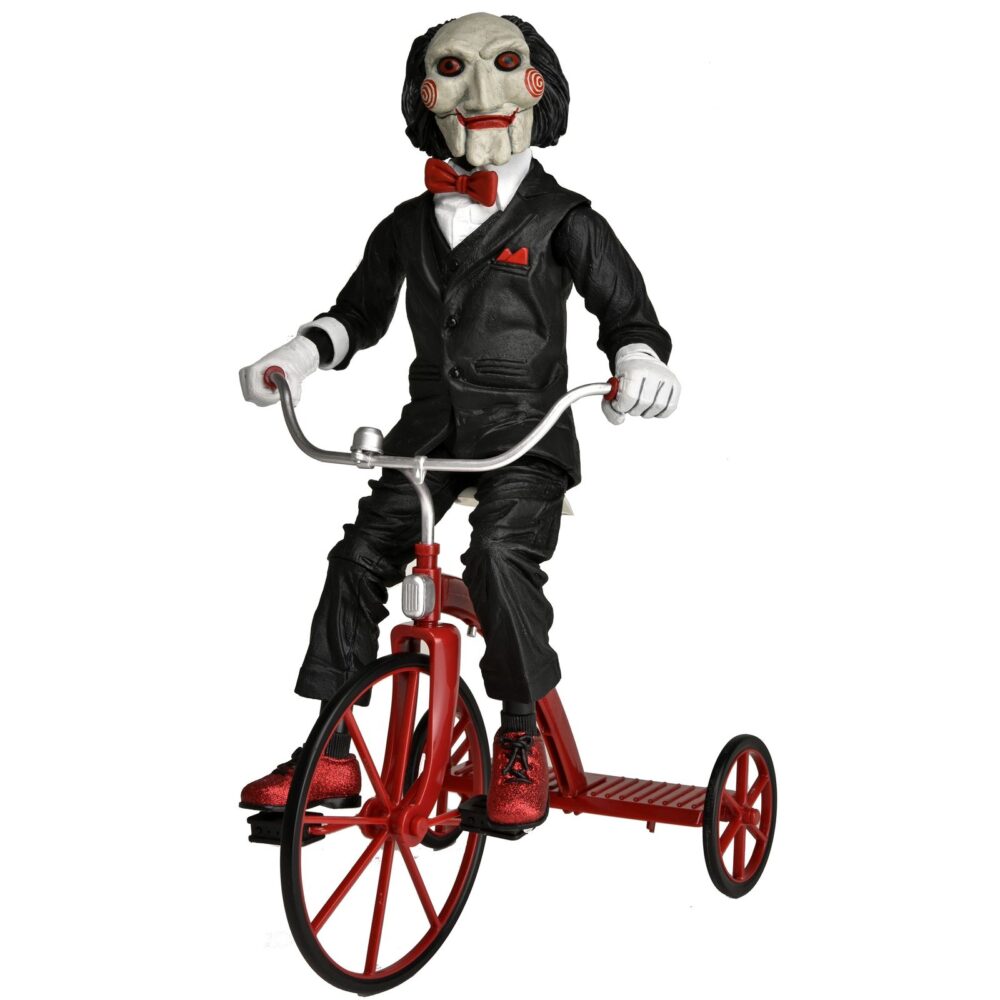 BILLY THE PUPPET ON TRICYCLE SAW 12″. Neca reincorpora en su catálogo esta figura de 30 cm de Billy the Puppet, el siniestro muñeco de la saga "Saw". Se muestra montado en su icónico triciclo rojo con zapatos de purpurina e incluye función de sonido con frases de la película. Emplea 3 baterías de 1.5V LR44 (incluidas). Se presenta en una caja con ventana.