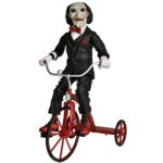 BILLY THE PUPPET ON TRICYCLE SAW 12″. Neca reincorpora en su catálogo esta figura de 30 cm de Billy the Puppet, el siniestro muñeco de la saga "Saw". Se muestra montado en su icónico triciclo rojo con zapatos de purpurina e incluye función de sonido con frases de la película. Emplea 3 baterías de 1.5V LR44 (incluidas). Se presenta en una caja con ventana.