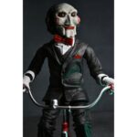 BILLY THE PUPPET ON TRICYCLE SAW 12″. Neca reincorpora en su catálogo esta figura de 30 cm de Billy the Puppet, el siniestro muñeco de la saga "Saw". Se muestra montado en su icónico triciclo rojo con zapatos de purpurina e incluye función de sonido con frases de la película. Emplea 3 baterías de 1.5V LR44 (incluidas). Se presenta en una caja con ventana.