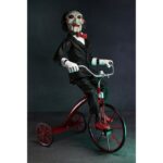 BILLY THE PUPPET ON TRICYCLE SAW 12″. Neca reincorpora en su catálogo esta figura de 30 cm de Billy the Puppet, el siniestro muñeco de la saga "Saw". Se muestra montado en su icónico triciclo rojo con zapatos de purpurina e incluye función de sonido con frases de la película. Emplea 3 baterías de 1.5V LR44 (incluidas). Se presenta en una caja con ventana.